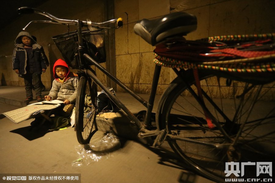 2015年11月8日晚，鄭州，7歲男孩涵洞橋下陪父母練攤，路邊燈下坐小馬扎上寫作業(yè)。