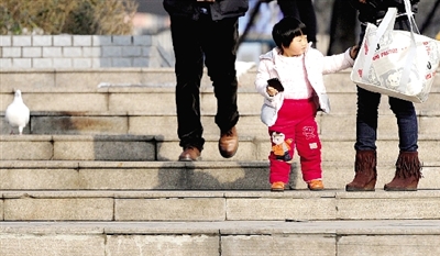昨天中午，一女孩拉著家長的手在紫荊山公園曬太陽，氣溫升高，孩子的棉衣解開了扣子。